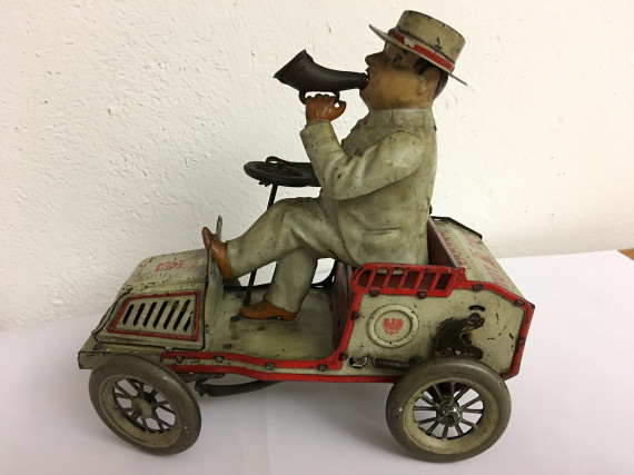 Blechspielzeug - Mann mit Hut sitzt lässig im offenen Auto. Ein Fuß auf dem Amaturenbrett, mit der linken Hand hat er eine Tute an den Mund geführt.
